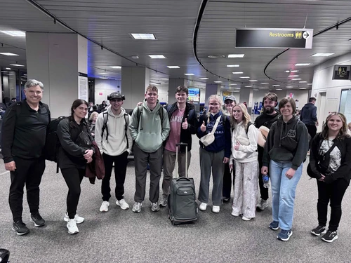 A group of travelers standing together in an airport terminal, smiling at the camera with luggage and backpacks.