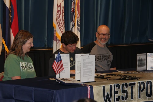 This image depicts a young man between his parents signing a paper. His parents are smiling at him. On the table is a table cloth that days West Point. Behind them are three flags. 