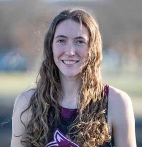This image depicts a girl in a track uniform smiling at the camera with the background blurred. The girl has wavy dirty blonde hair. 