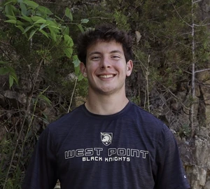 This image depicts a young man smiling in front of a camera for a headshot. Behind him are trees. He is wearing a grey shirt that says "West Point Black Knights". 