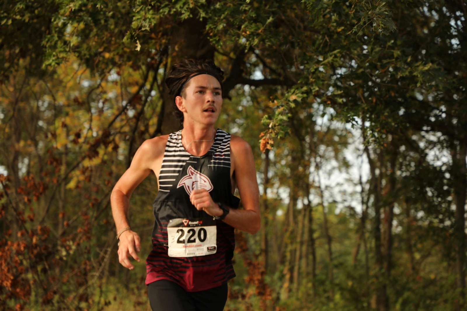 This image depicts a college aged man running through a forested area in a track uniform. 