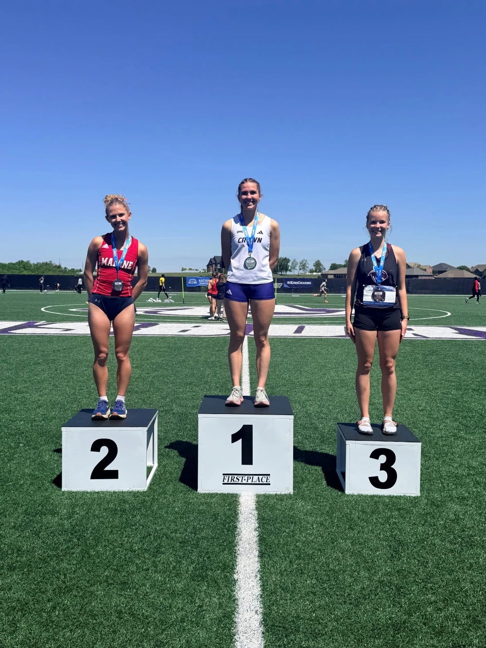 This image depicts three girls standing on podiums ranked from 3rd place to 1st. They are wearing track uniforms. 