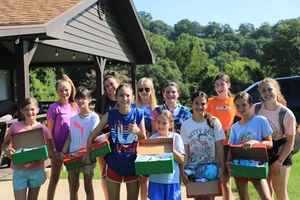 a group of of ten girls around the age of 10 posing for a picture outside of a cabin with their two camp counselors. 