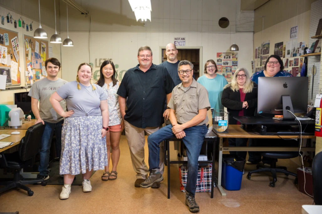 Nine adults pose for a group photo in a classroom filled with colorful artwork, bulletin boards and worktables. Eight stand while one man sits on a stool near the center. Most are smiling at the camera, and a large computer monitor sits on the table to the right.