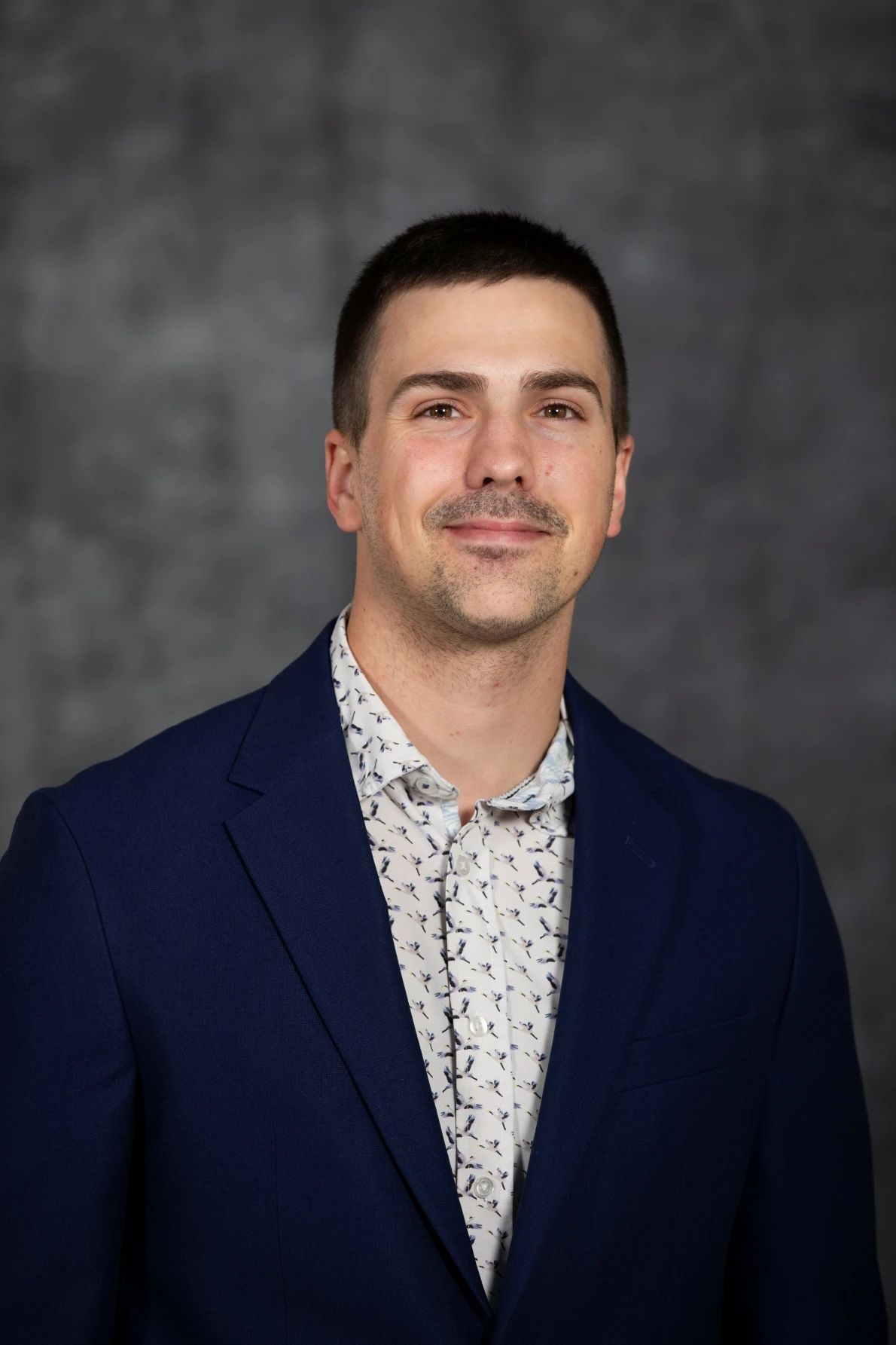 This image depicts a man smiling in front of a grew background. 