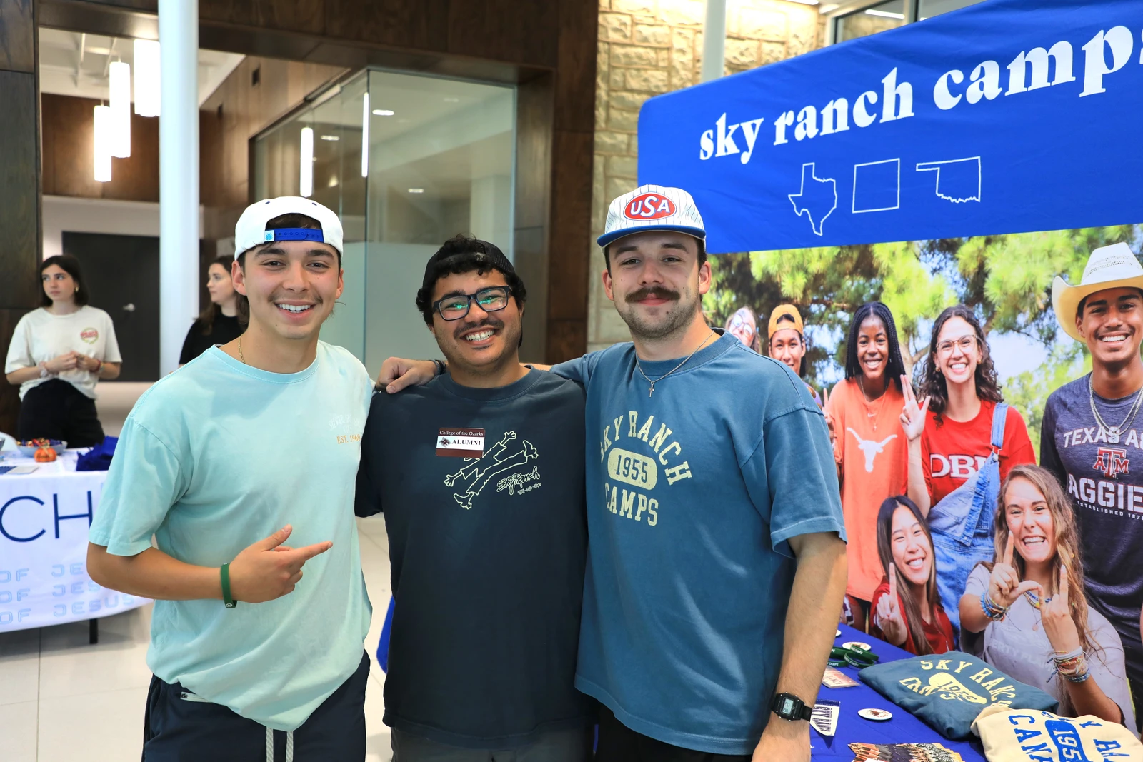 This image depicts three men with their arms around each other smiling at the camera. The words, "Sky Ranch Camps" is in the background. 