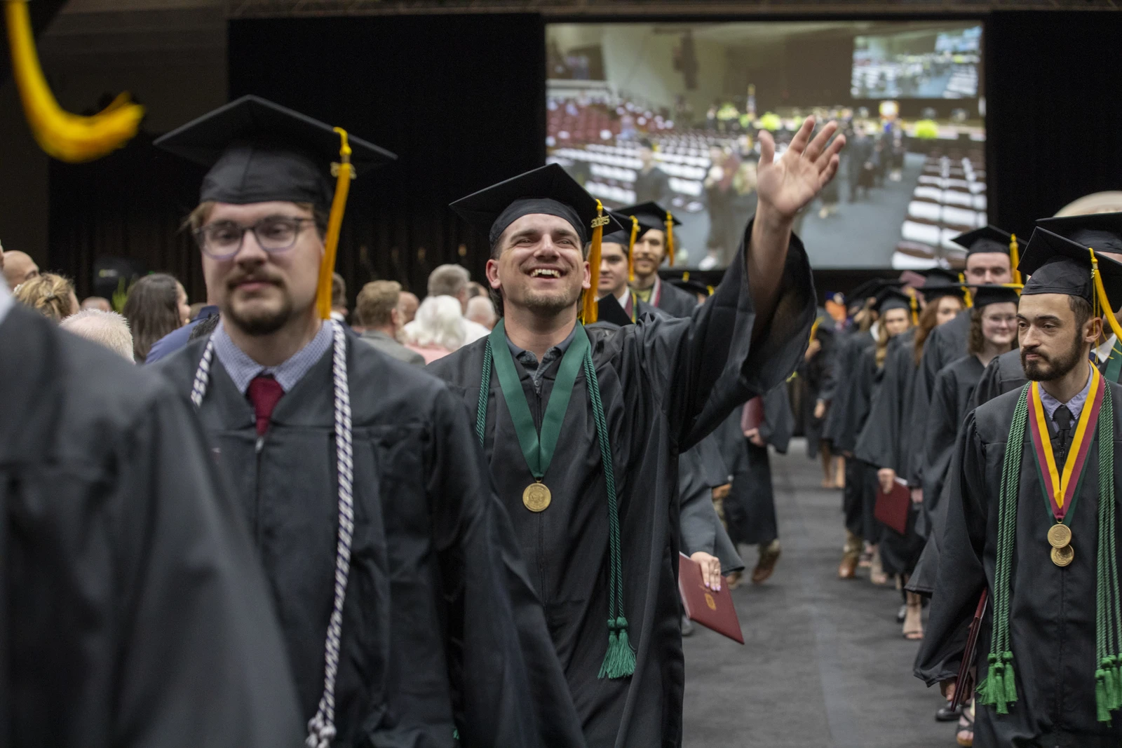 A group of college graduates in black caps and gowns walks in a procession through a crowded auditorium. At the center, a smiling graduate wearing glasses and a green honor cord raises his right hand to wave to the audience. A gold medal hangs around his neck, and he carries a red diploma cover in his left hand. Other graduates walk alongside him, wearing green cords and gold medals, while family members and guests sit nearby. A large screen in the background displays a live view of the ceremony.