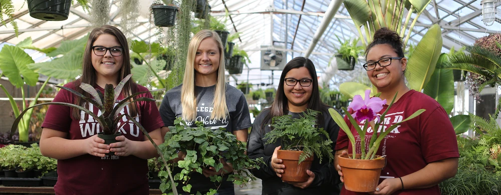 Four woman holding various potted plants in a greenhouse.
