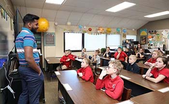 College student in polo and jeans in front of classroom of fourth graders dressed in red.